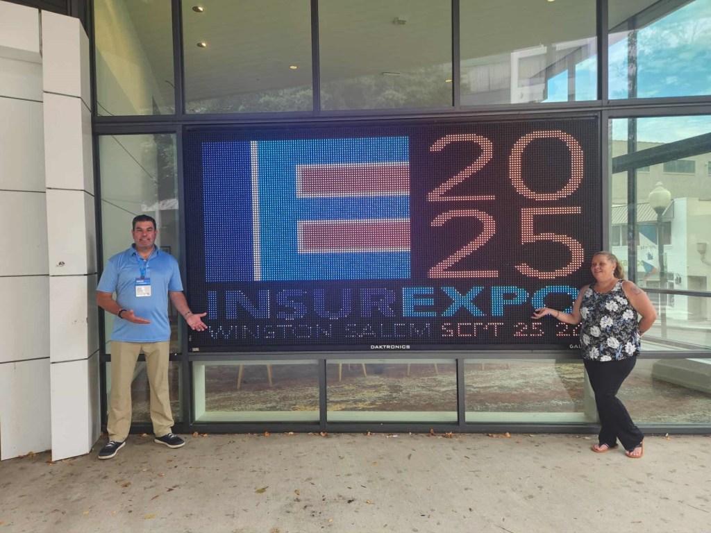Two individuals posing in front of a large digital sign displaying the text 'INSUREXPO 2025 WINSTON SALEM SEPT 25' outside a building.